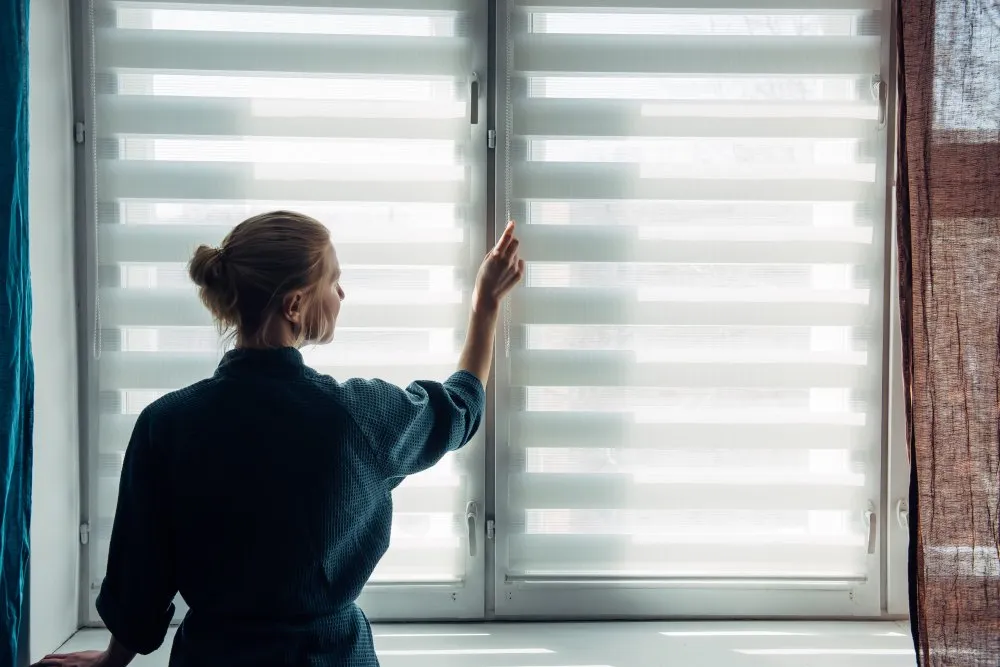 Store fenêtre Femme ajustant un store sur une fenêtre pour réguler la luminosité intérieure d’une pièce, en journée.
