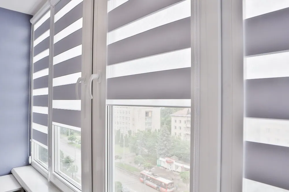 Stores gris Stores gris installés sur des fenêtres en PVC blanches dans un intérieur lumineux avec vue sur des immeubles résidentiels et une avenue urbaine.