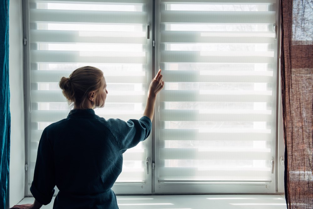 Femme ajustant un store sur une fenêtre pour réguler la luminosité intérieure d’une pièce, en journée.