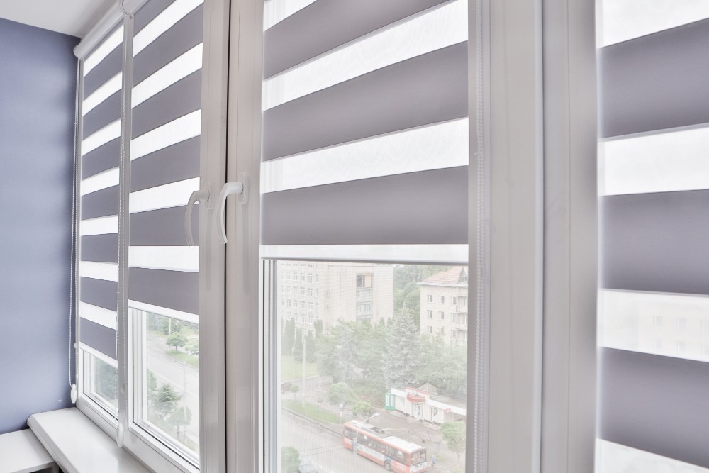 Stores gris installés sur des fenêtres en PVC blanches dans un intérieur lumineux avec vue sur des immeubles résidentiels et une avenue urbaine.